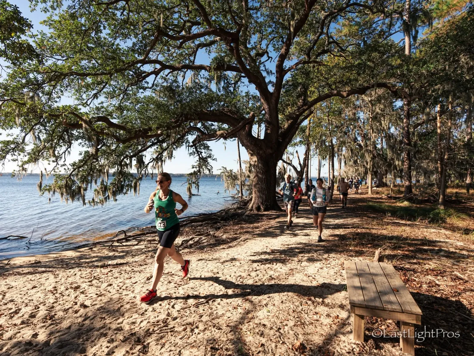 A female runner in a green tank top and black shorts runs along a sandy path next to a body of water, with other runners visible in the background under large, moss-draped trees.