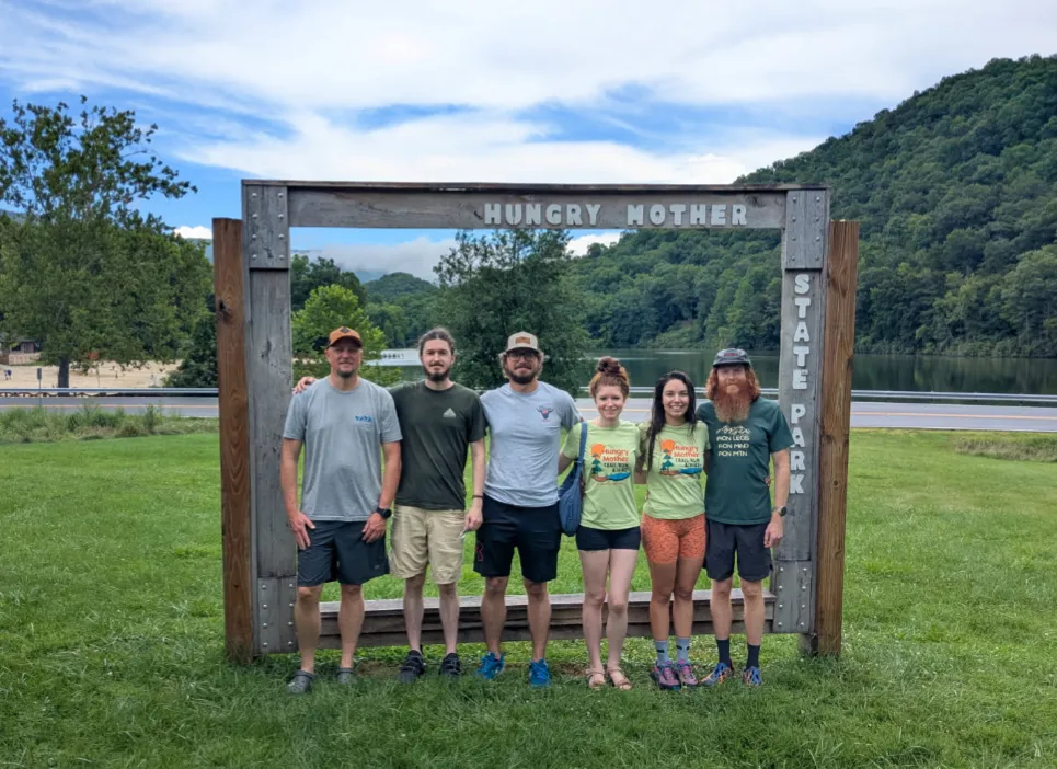 A group of six people stand in front of a wooden sign that reads "Hungry Mother State Park" with a lake and forested hills in the background.