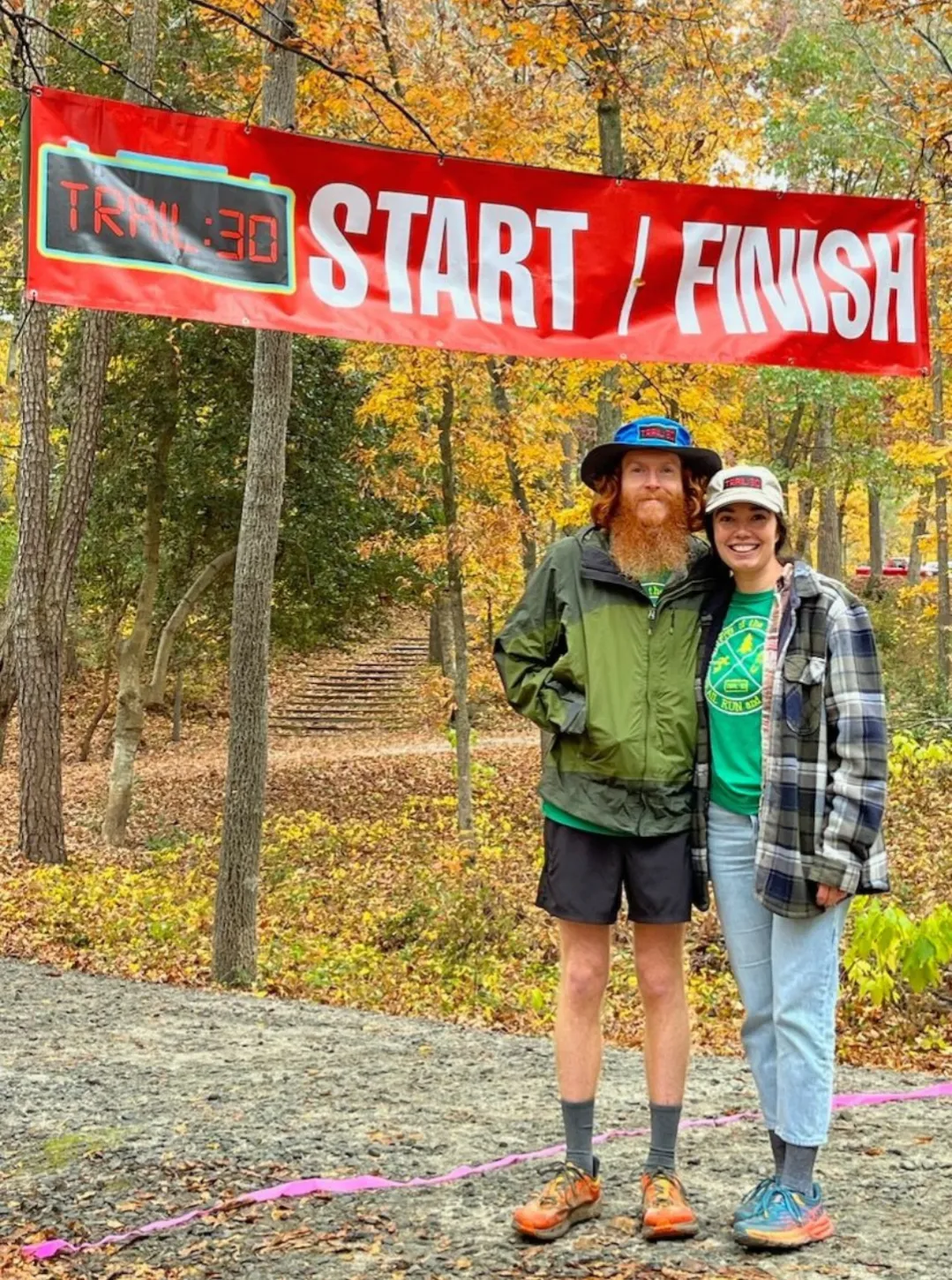 Two people smiling in front of a red start/finish banner in a forest with autumn foliage.