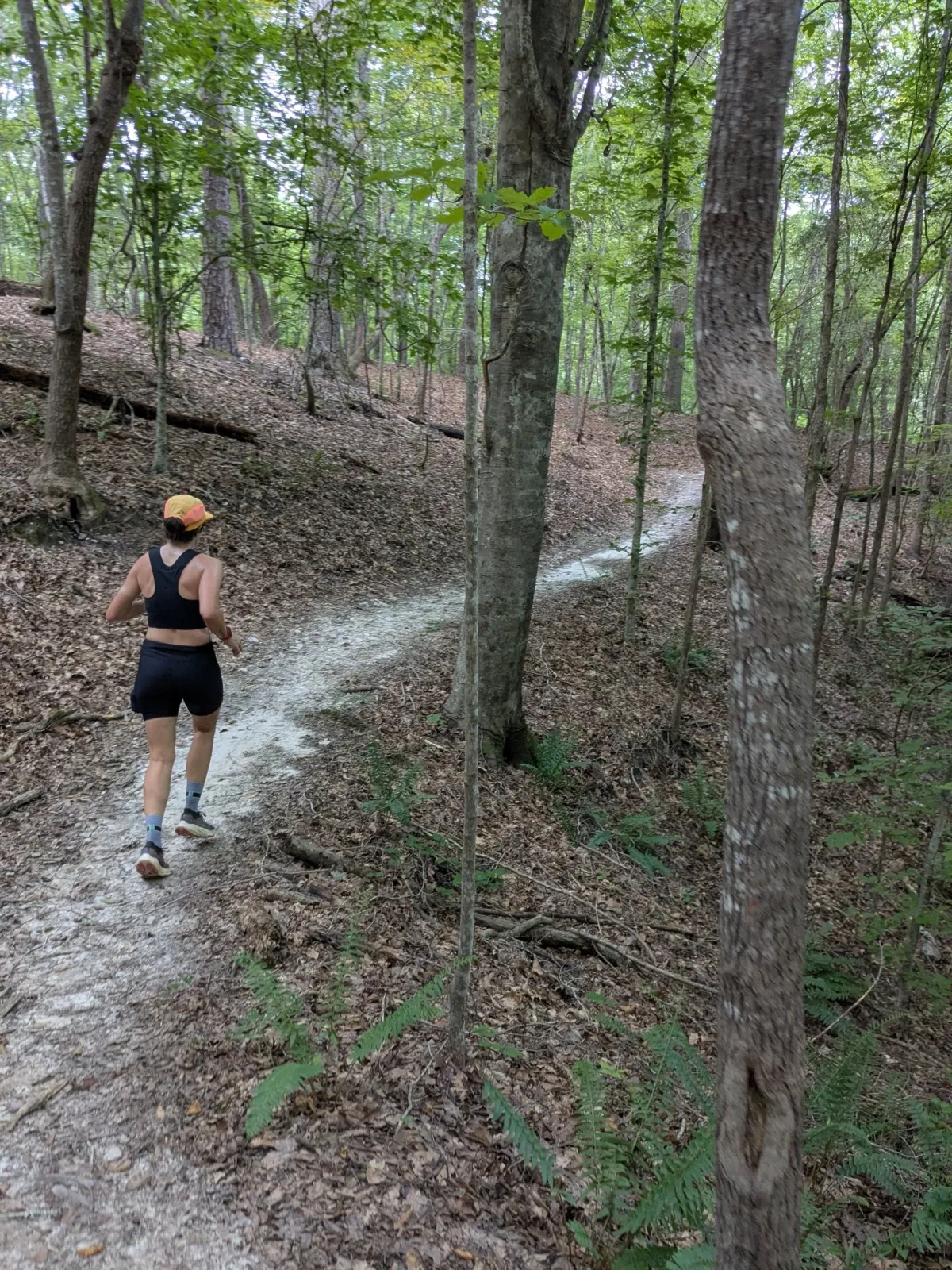 A female runner with a yellow hat and black athletic wear runs on a dirt trail through a forest.