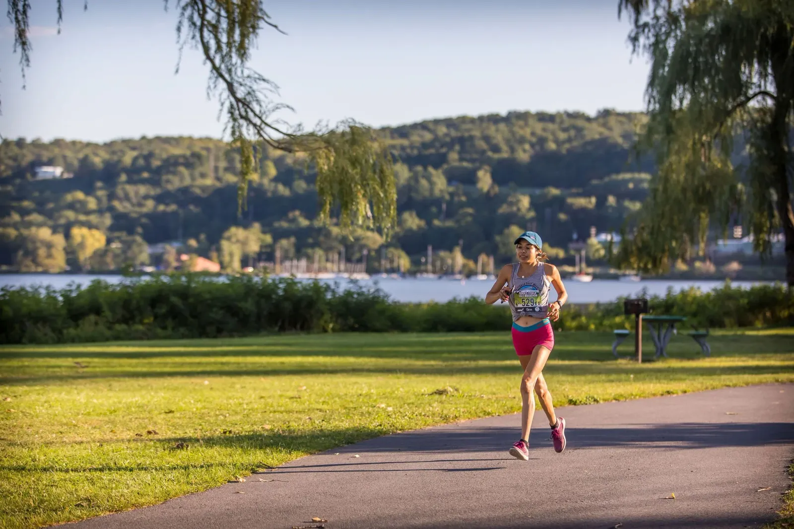 A female runner with a colorful race shirt runs along a paved path with a waterfront and tree-lined hills in the background.