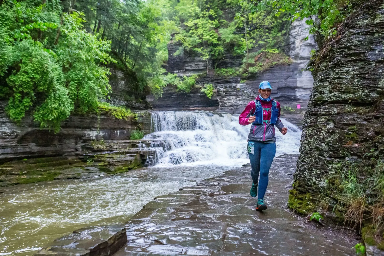 A female runner with a blue hat and pink jacket runs along a wet stone path next to a waterfall.