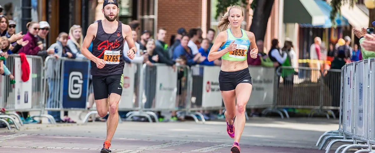 A male and female runner are shown in the foreground, running towards the camera during a race. Spectators are visible behind barriers in the background.