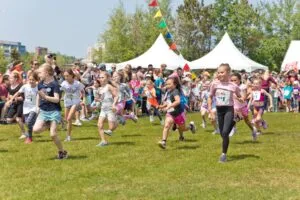A group of young girls in athletic wear running on a grassy field, with tents and flags in the background, at the start of a race.