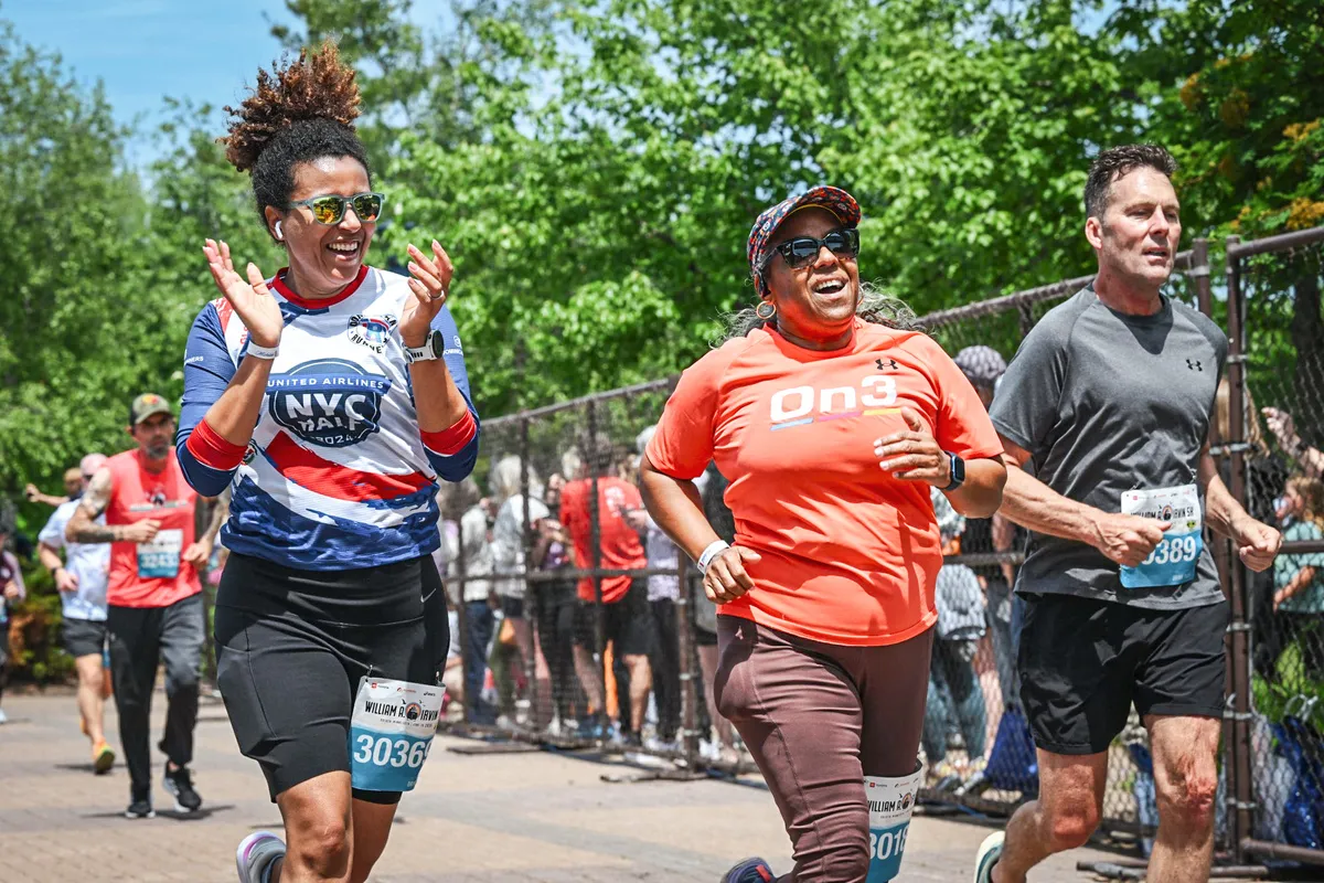 Three runners smiling and running on a road, with trees and a fence in the background.
