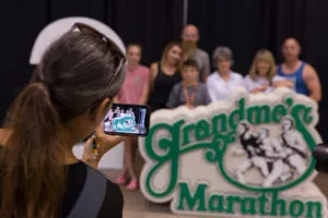 A group of people, including children and adults, pose for a photo in front of a large 'Grandma's Marathon' sign.