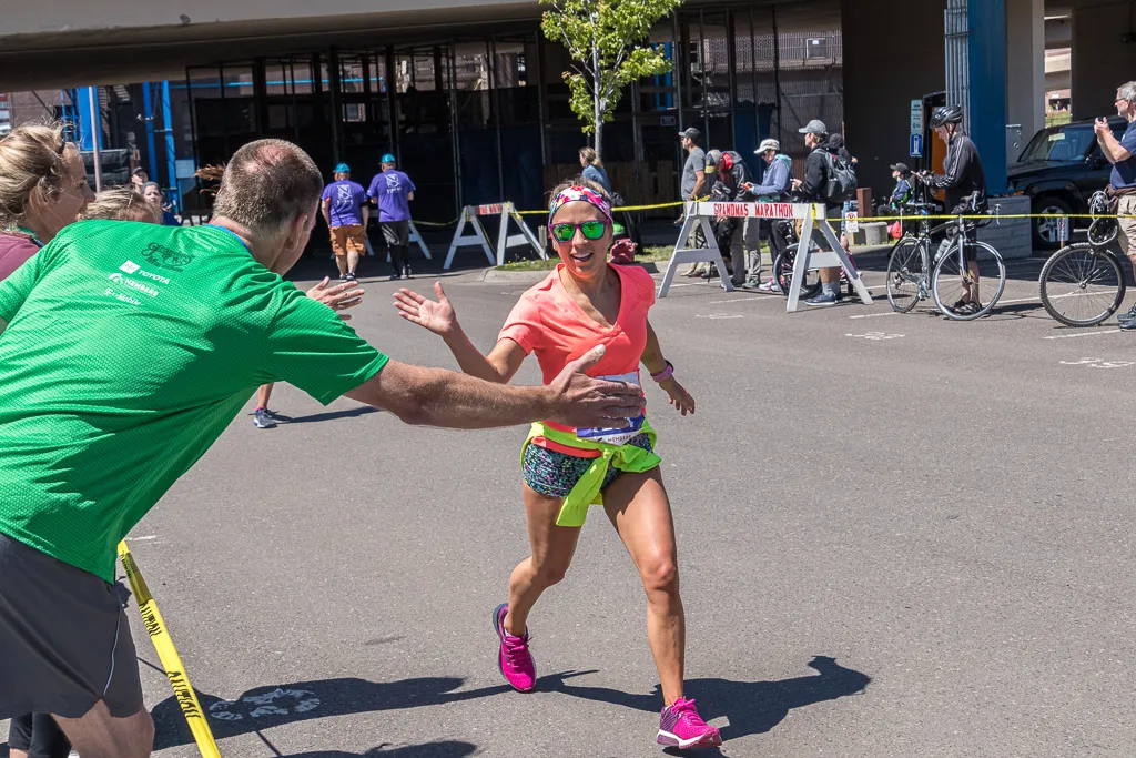 A female runner in a pink shirt and colorful shorts gives a high-five to a volunteer in a green shirt during Grandma's Marathon.