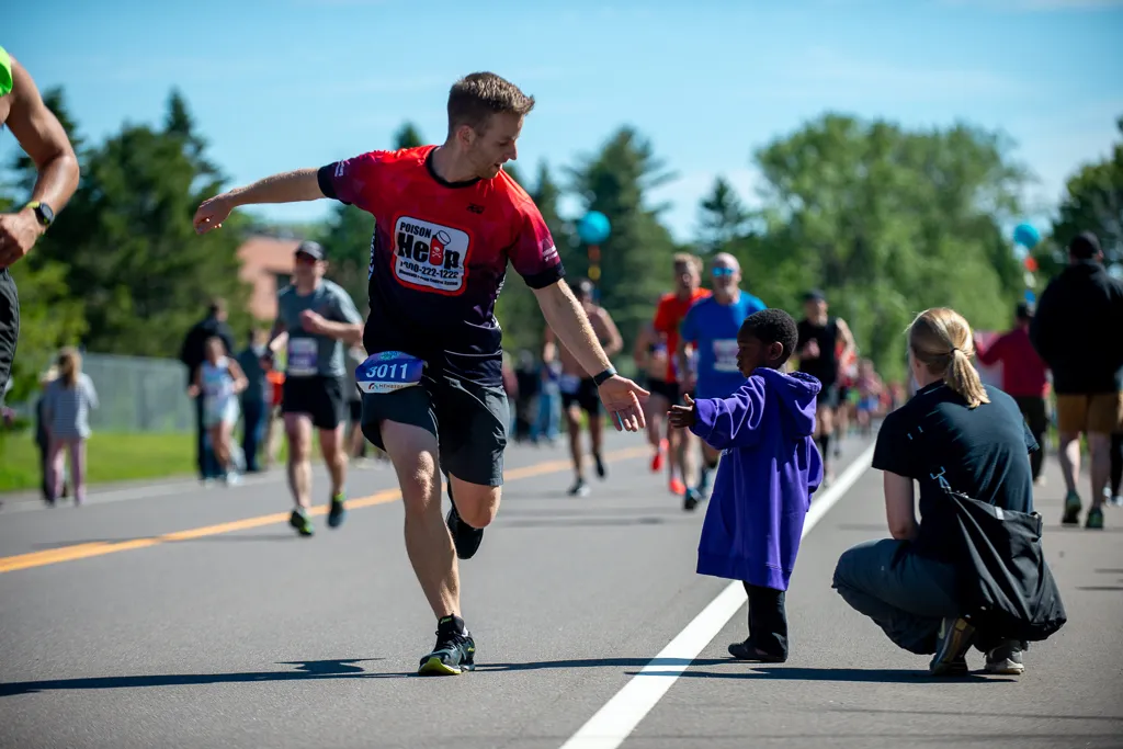 A male runner in a red and black shirt high-fives a young child in a purple hoodie on a race course.
