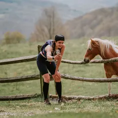 A female runner pauses on a trail to take a selfie with a horse standing behind a wooden fence.