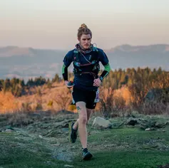 A runner with a bun runs on a grassy trail with mountains in the background at sunset.