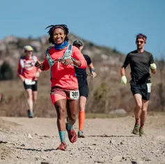 A female runner smiles as she runs on a dirt path, with other runners behind her and mountains in the background.