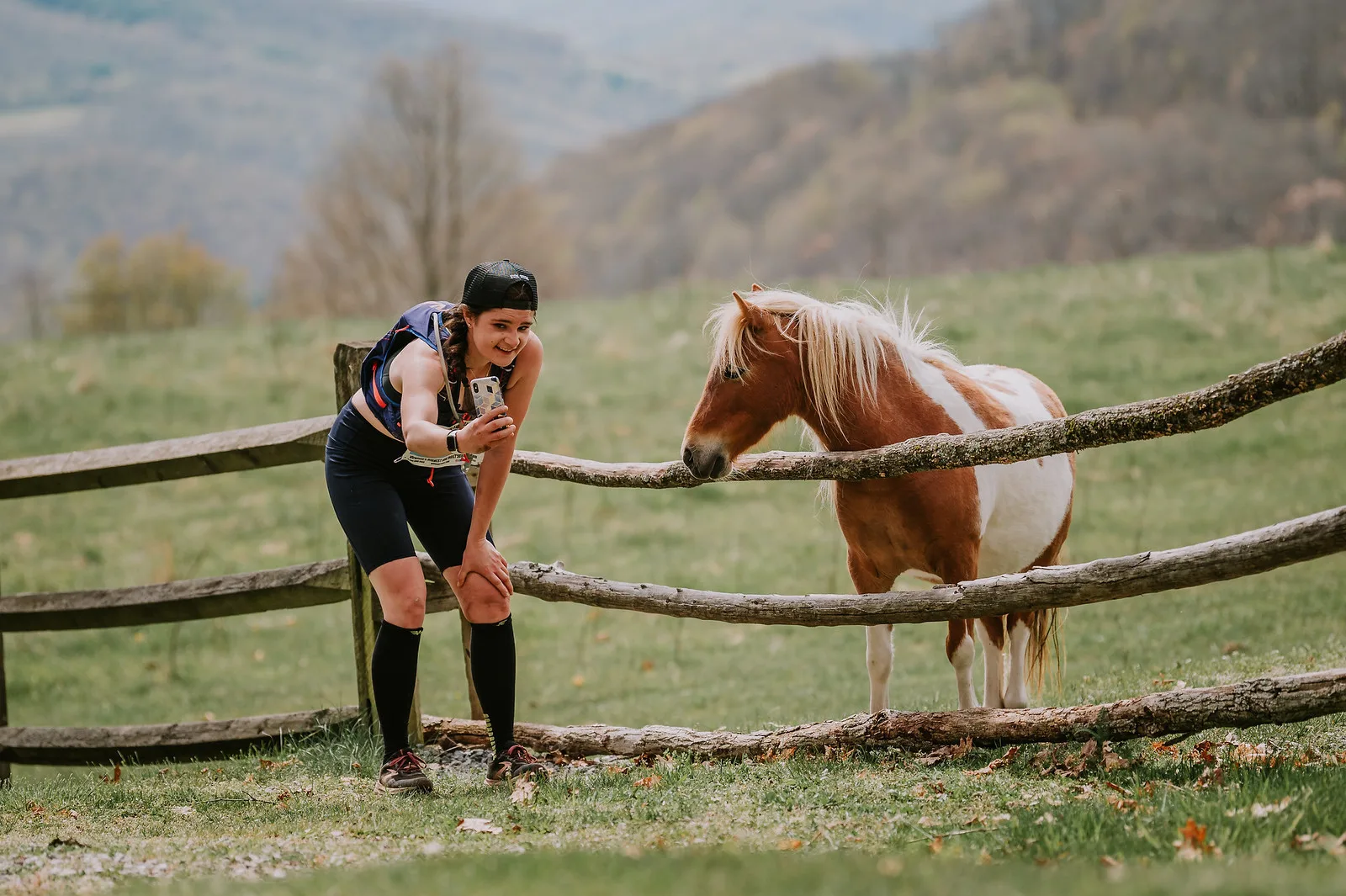 A female runner in athletic gear pauses on a trail to take a photo of a horse standing behind a wooden fence.
