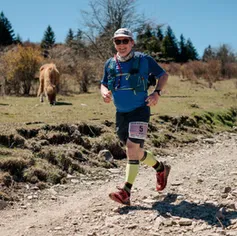 A male runner wearing a blue shirt, black shorts, and yellow compression socks runs on a dirt trail. A Highland cow grazes in the background.