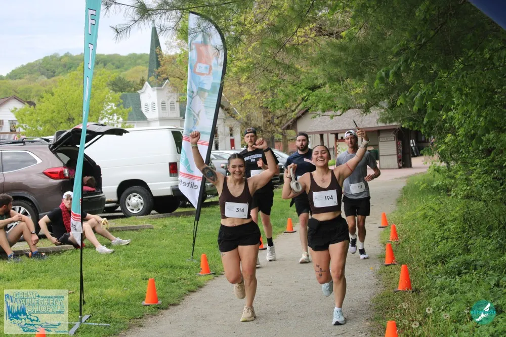 Two female runners with their arms raised in celebration as they cross the finish line, with other runners and spectators in the background.