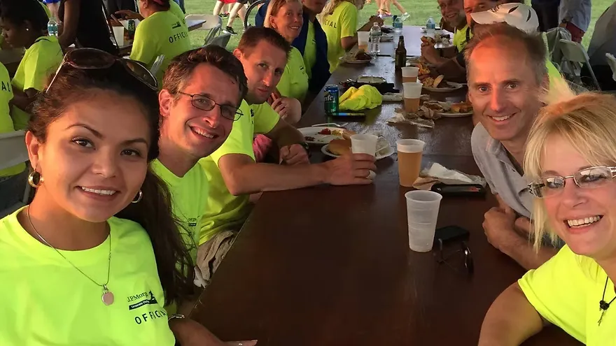 A group of people, some wearing bright yellow shirts with 'OFFICIAL' visible, smiling and sitting at a long table, suggesting a post-race gathering or event.