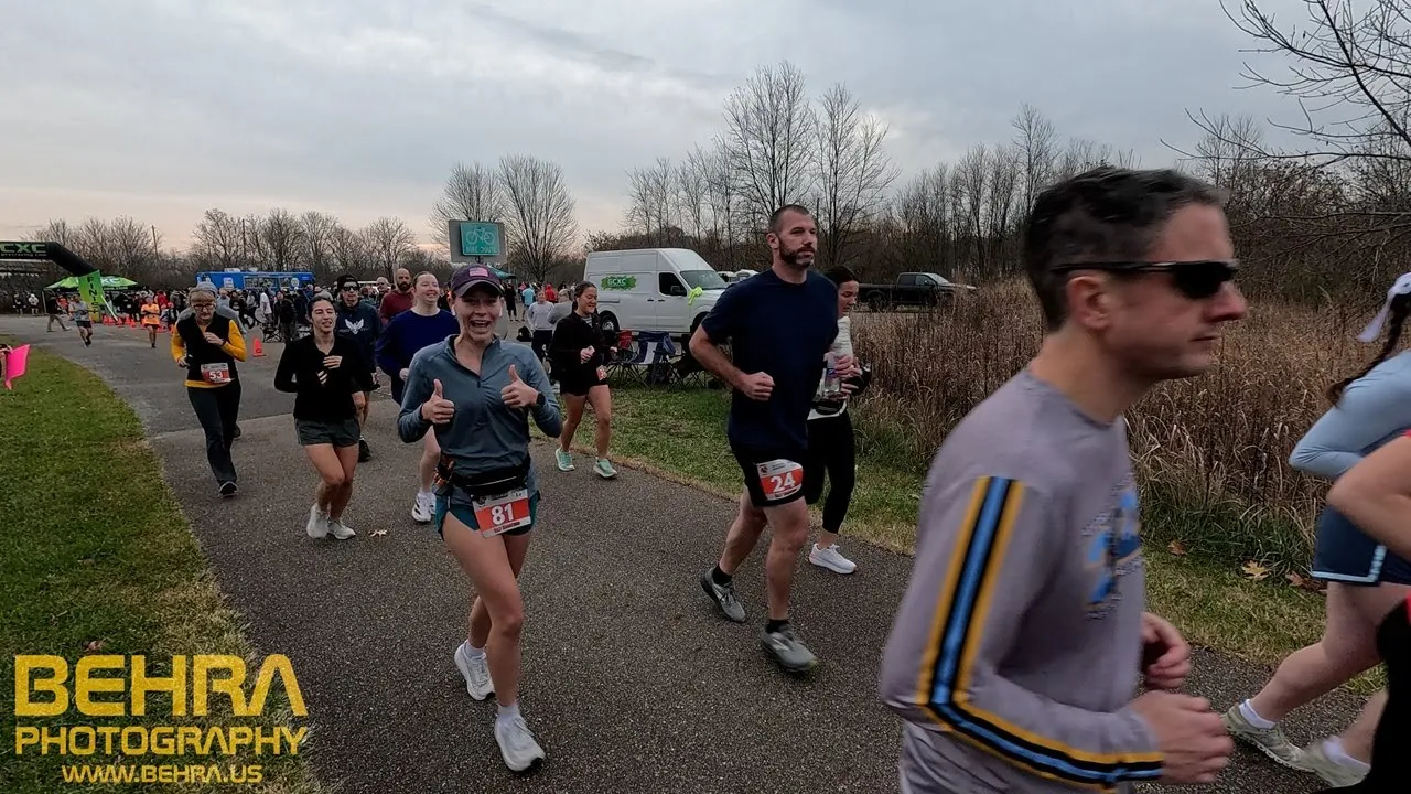 Runners smiling and giving thumbs up at the start of a race, with a crowd in the background.