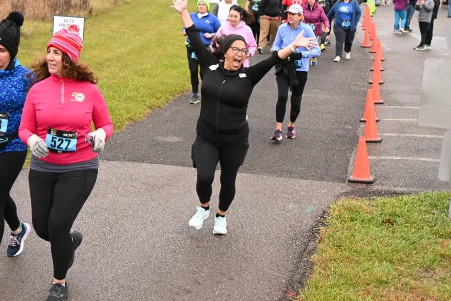 A group of female runners on a paved path, with one runner in the foreground in a black outfit with her arms raised in celebration and a wide smile.