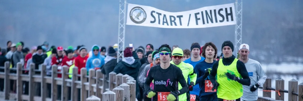 Runners lined up at the start/finish line of a race in cold weather, with a banner overhead.