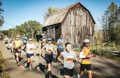 Runners race past a rustic wooden barn on a sunny day.