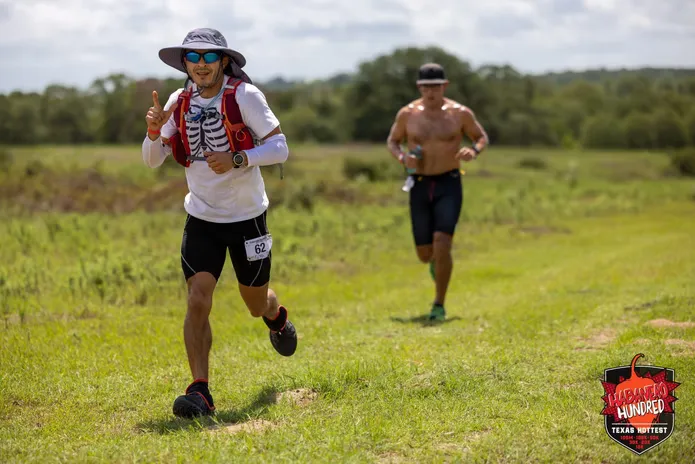 A runner wearing a hat and sunglasses, with a bib number 62, runs on a grassy course with another runner in the background. A race logo is visible in the bottom right corner.