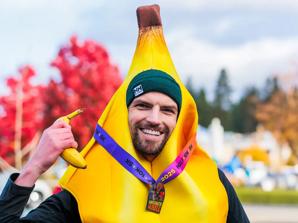 A smiling runner dressed as a banana, wearing a green beanie and a medal from the Halloween Hustle race.