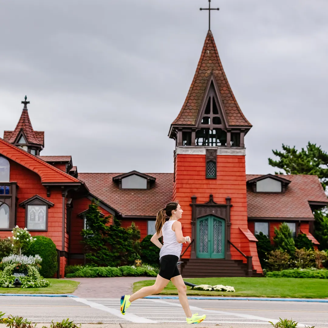 A female runner in a white tank top and black shorts runs across a crosswalk with a distinctive red church building in the background.