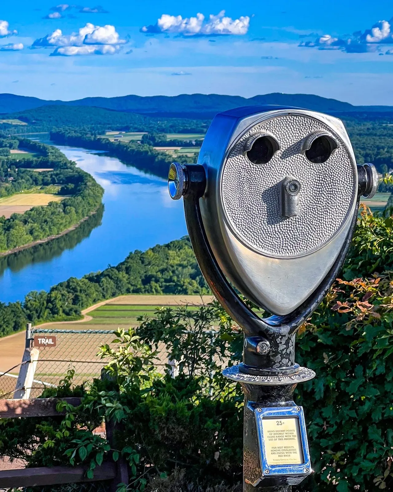 Scenic view of a winding river with lush green trees and rolling hills in the background, with a coin-operated viewer in the foreground and a sign indicating a trail.