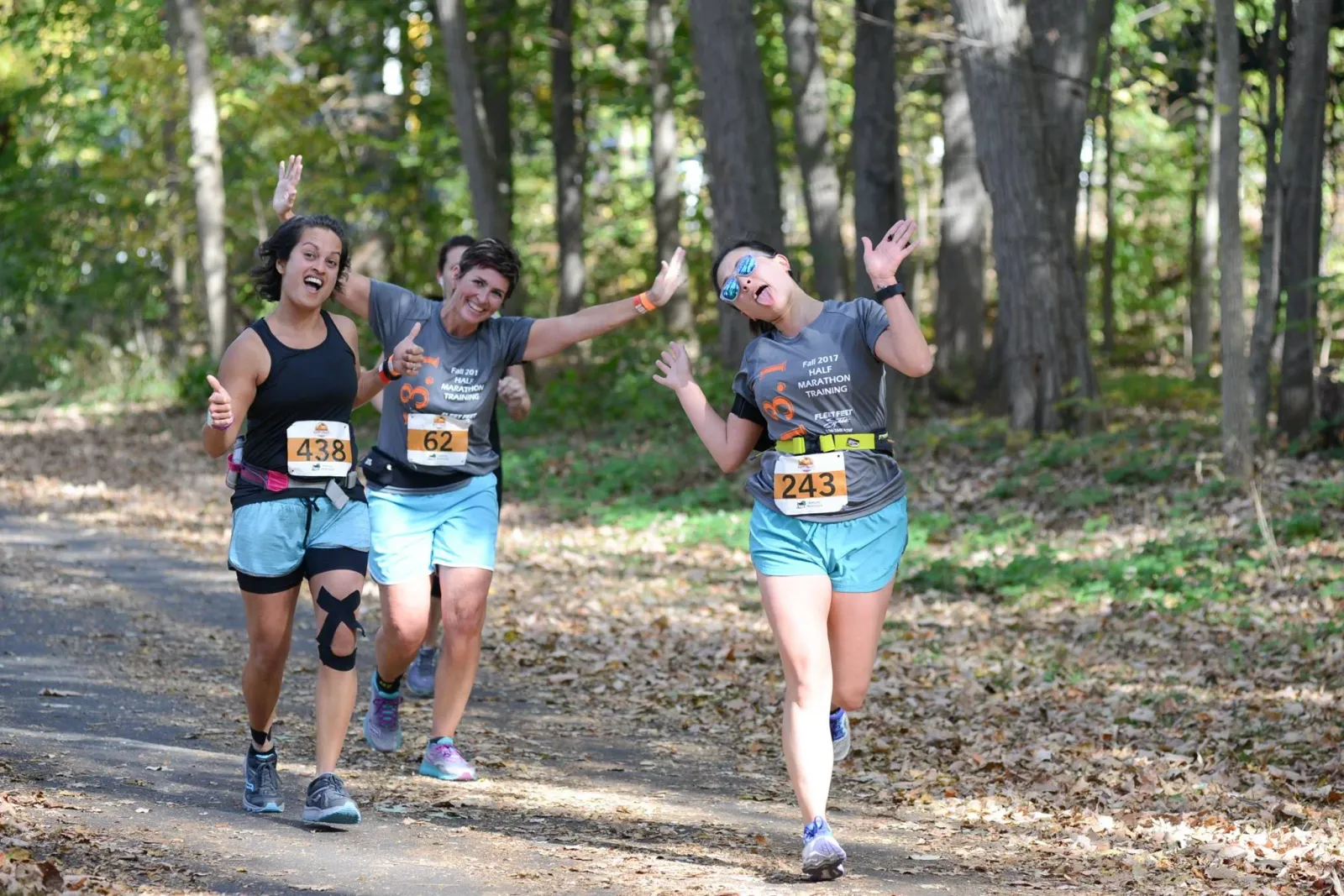 Three women running on a wooded path, two giving thumbs up and one sticking her tongue out, all wearing race bibs.