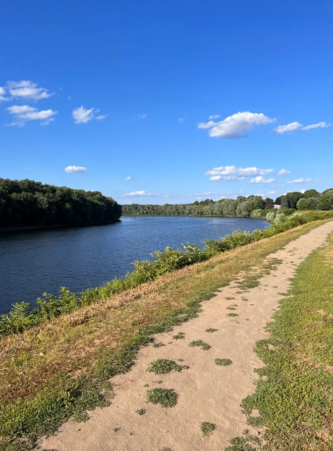A dirt path runs alongside a calm body of water, with lush green trees lining the opposite bank under a clear blue sky with scattered clouds.
