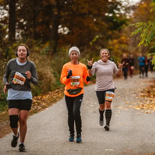 Three runners on a paved path surrounded by trees with autumn foliage, two in the foreground smiling and one making a rock-and-roll hand gesture.