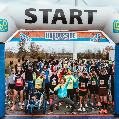 A large group of runners cheer and raise their hands at the start line under an inflatable archway for the Harborside Half Marathon & 5K.