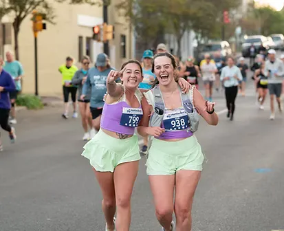 Two female runners smiling and laughing, with one giving a peace sign, during a road race.