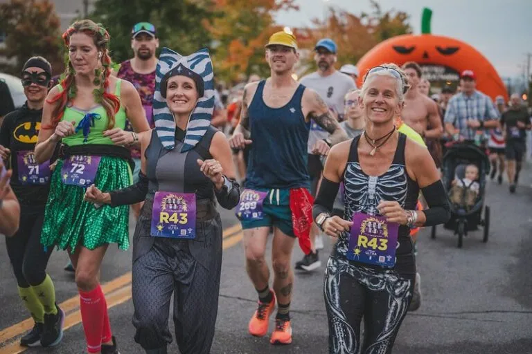 Runners in Halloween costumes, including a Batman-themed runner, a Poison Ivy-themed runner, and a Star Wars Ahsoka Tano-themed runner, participating in a race with a large pumpkin arch in the background.