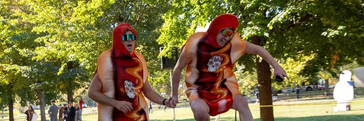 Two people in hot dog costumes with sunglasses running and holding hands, with trees and other runners in the background.