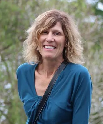 A smiling woman with blonde, wavy hair and a teal shirt, with a black strap across her shoulder.