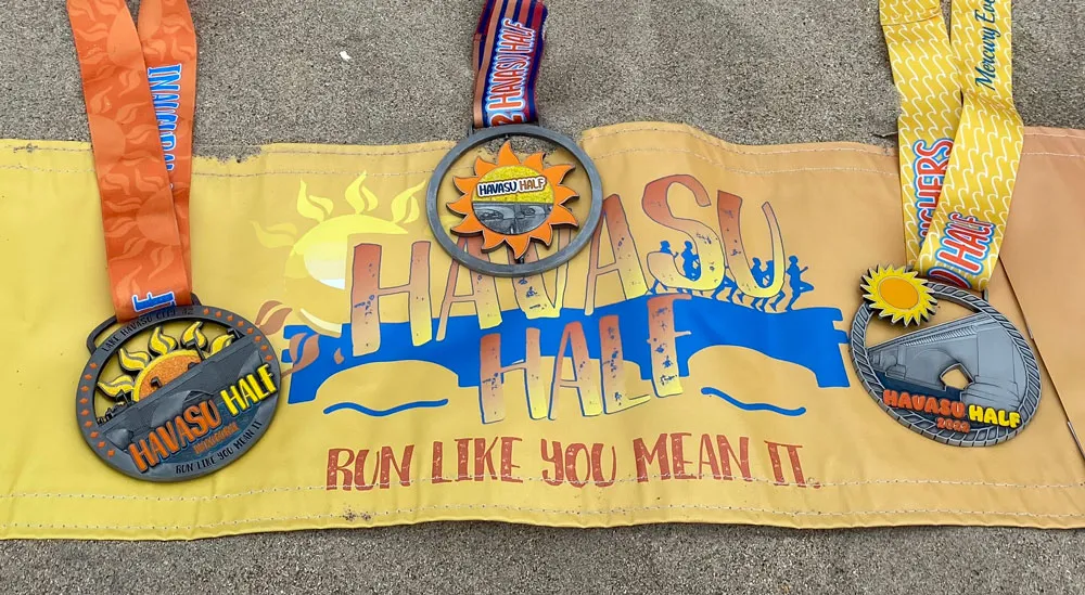 Three Havasu Half Marathon medals displayed on a yellow banner with the race logo and slogan 'Run Like You Mean It', laid out on sand.