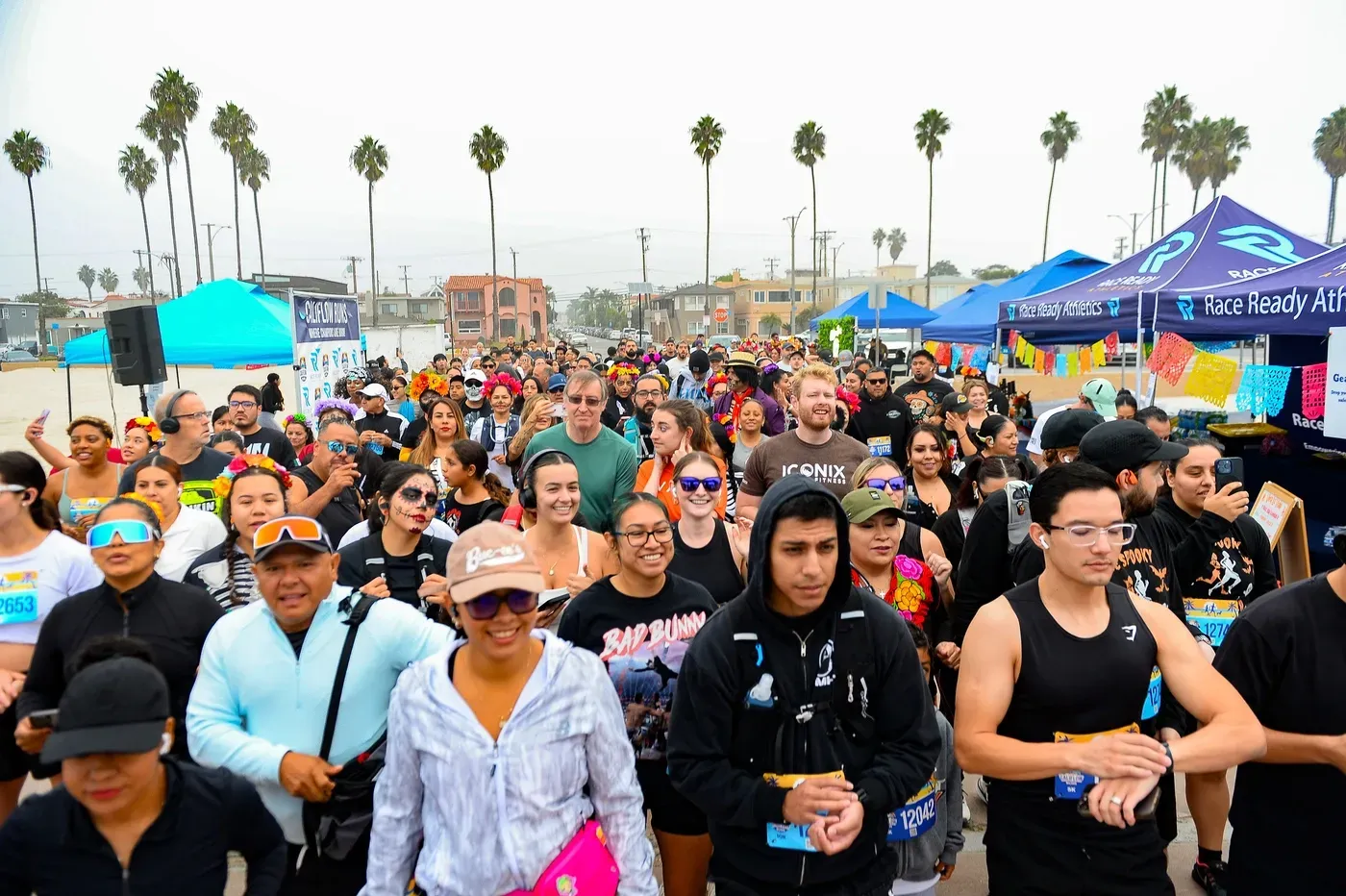 A large crowd of diverse runners, many wearing festive attire and face paint, gather at the start of a race on a paved path near a beach, with palm trees and tents in the background.