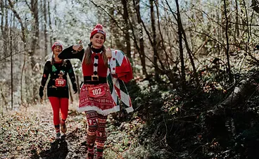 Two women in festive holiday running attire, one giving a thumbs up, running on a wooded trail.