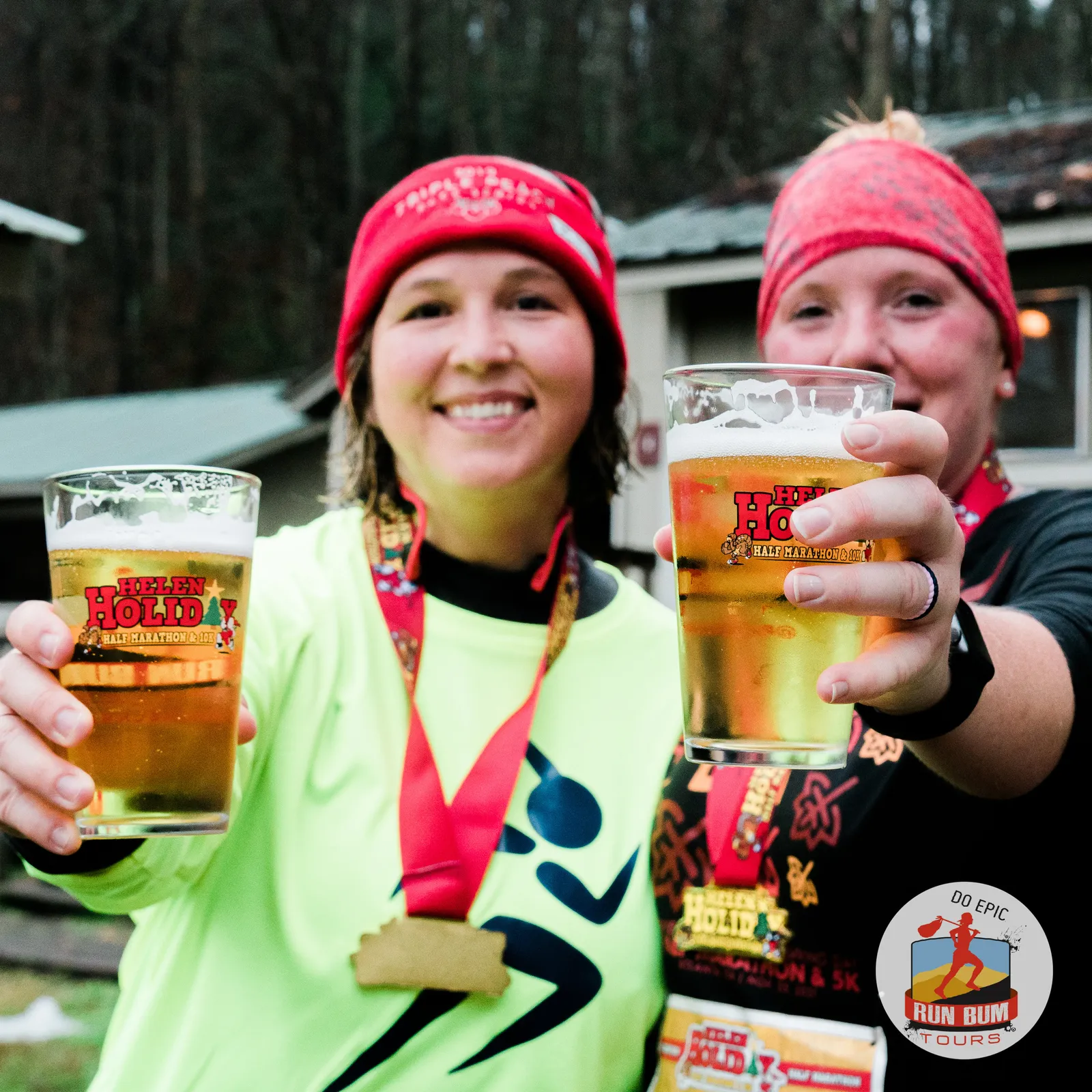 Two smiling runners wearing medals and race beanies hold up glasses of beer with the 'Helen Holiday Half Marathon & 10K' logo.