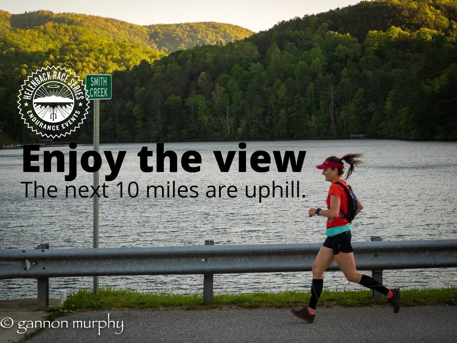 A female runner runs along a road next to a lake with forested hills in the background. Text overlay reads 'Enjoy the view. The next 10 miles are uphill.'