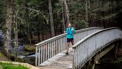 A male runner smiles and gives a thumbs up while running across a wooden bridge on a wooded trail.