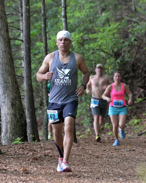 A male runner wearing a grey tank top with 'Atlanta Track Club' logo and bib number 140 runs on a wooded trail, with other runners behind him.