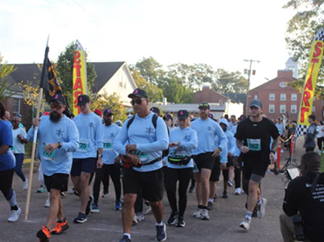 Runners gather at the start line of a race, with a checkered flag arch and yellow start banner visible.