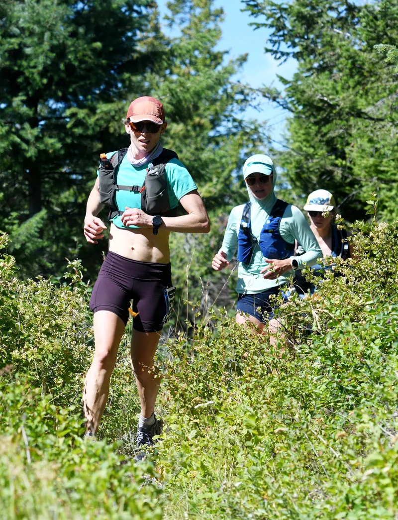 Three women running on a wooded trail, with the lead runner in focus.