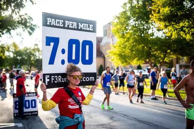 A volunteer holds a 'Pace Per Mile 7:00' sign at a road race, with runners in the background.