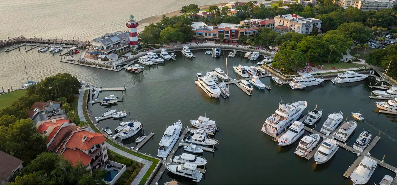 Aerial view of a harbor with numerous boats docked, a lighthouse, and buildings along the waterfront.