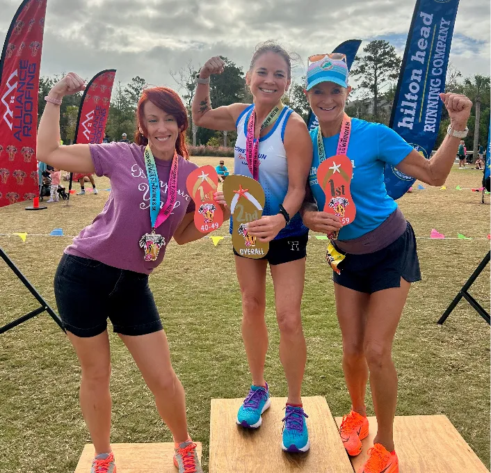 Three women posing on a podium, celebrating their race wins with medals and flip-flop awards.