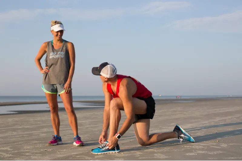 A man kneels on a sandy beach tying his running shoes while a woman stands nearby, both dressed in athletic wear.