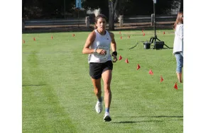 A female runner in a white tank top and black shorts runs towards the camera on a grassy field marked with small red flags.