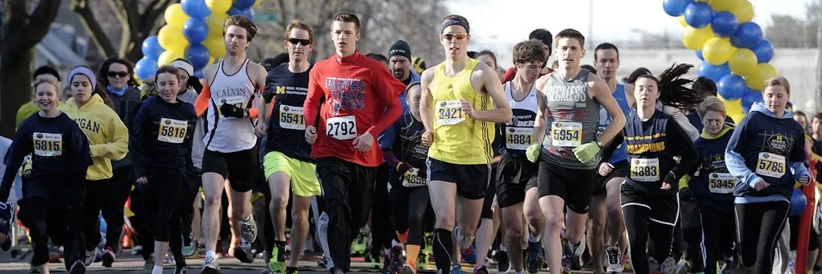 A group of runners, including men and women of various ages, are running forward at the start of a race. They are wearing race bibs and some are wearing team or event t-shirts. Yellow and blue balloons arch over the start line.